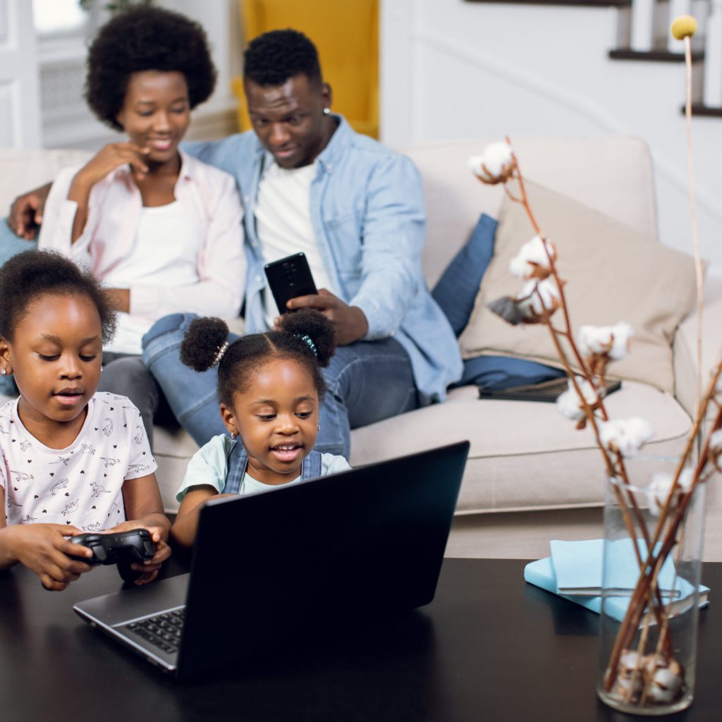 Cute african american sisters using laptop and joysticks for playing video games while their young parents resting on couch with smartphone in hands. Family at home.