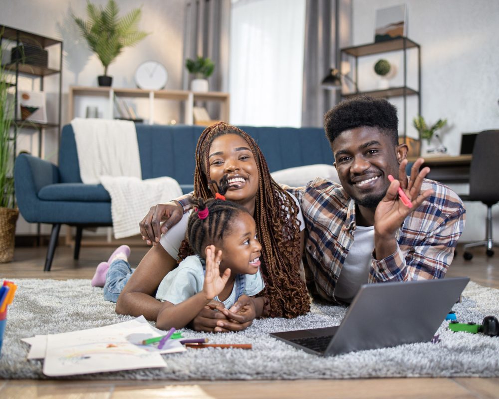 Positive young parents embracing pretty daughter while lying on floor with modern laptop, toys and pencils. Happy african family waving hands and smiling on camera.